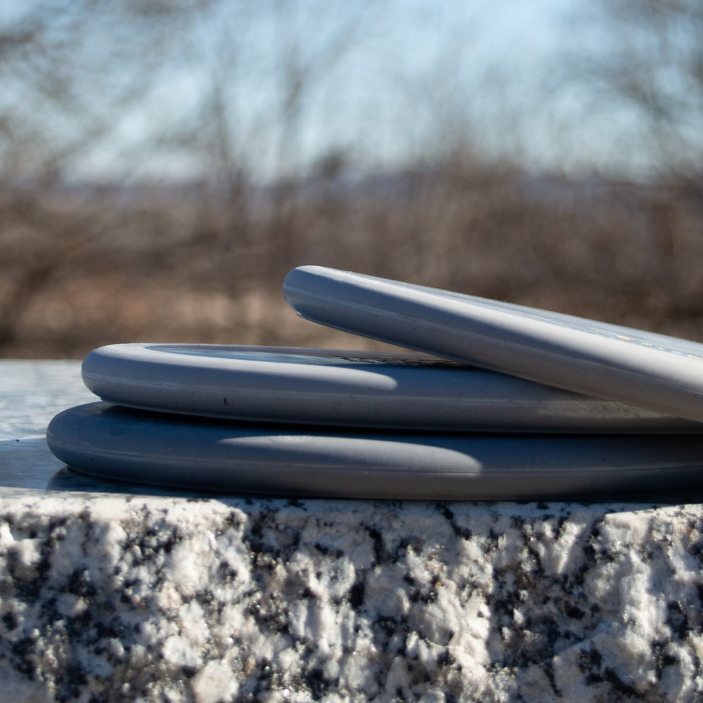 Three Chief - Rocky Lunar putters, gray and low profile with firm lunar plastic, are stacked on a textured stone outdoors against a blurred backdrop of leafless trees.