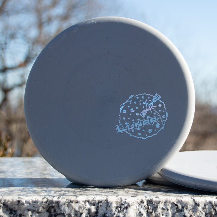 A gray Chief - Lunar Putt & Approach disc golf disc in durable Lunar plastic, featuring a "Lunar" logo with a guitar and moon design, rests on a granite surface outdoors.