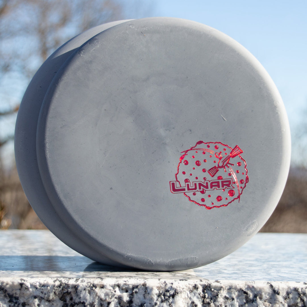 The Chief - Rocky Lunar disc golf disc, gray and made from firm lunar plastic, features a red "Lunar" logo and moon graphic, sitting on a granite outdoor surface.