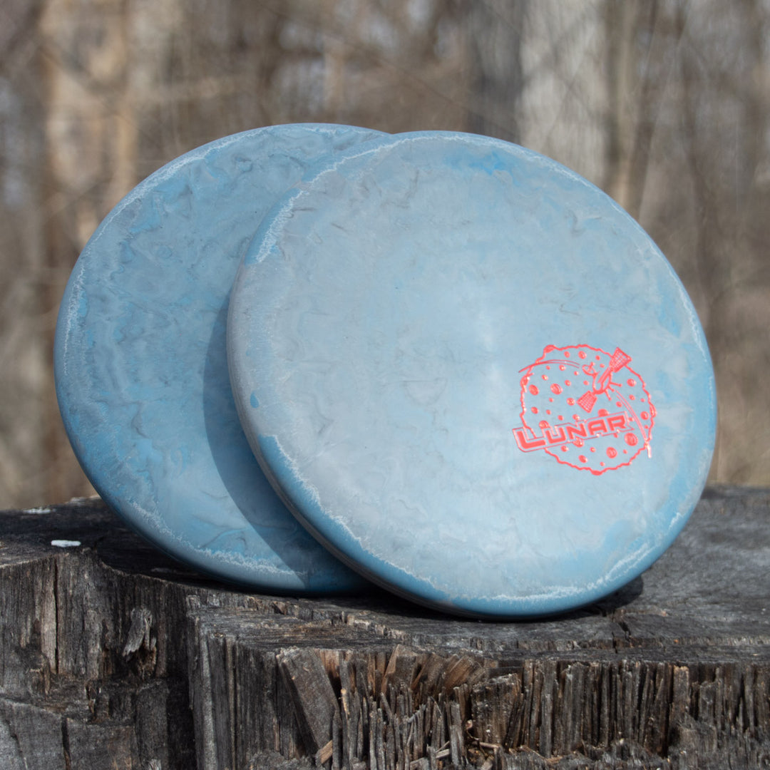 Two blue Magic - Lunar putters with red logos sit on a tree stump, ready for action. Renowned for their impressive glide and straight flights, they stand out against the blurred woodland background.