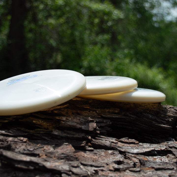 Three Magic - Diamond white putter discs, perfect for straight flights or beginners, are stacked on a rough tree trunk with softly blurred green foliage in the background.
