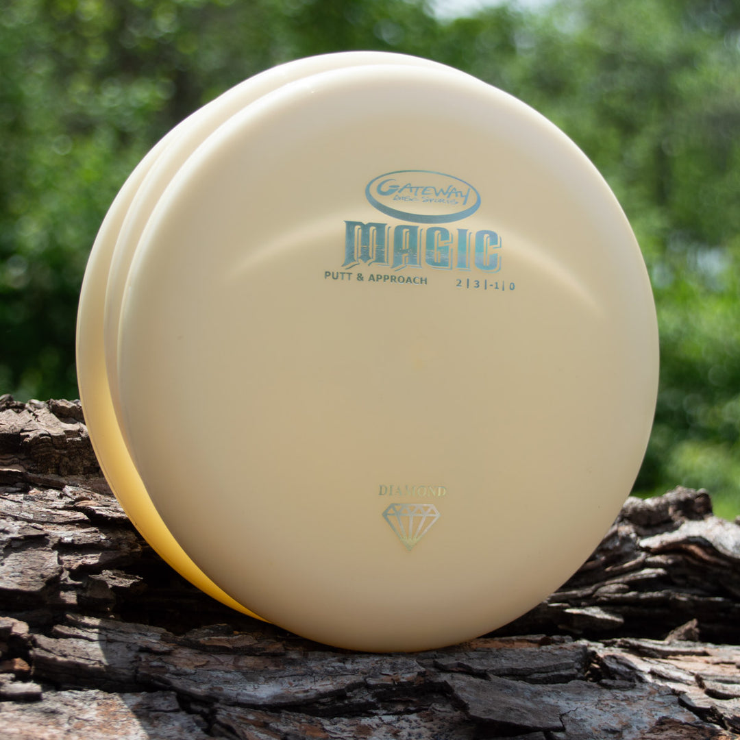 A pale yellow Gateway Magic - Diamond straight-flying putter disc stands upright on a tree log, surrounded by green foliage.