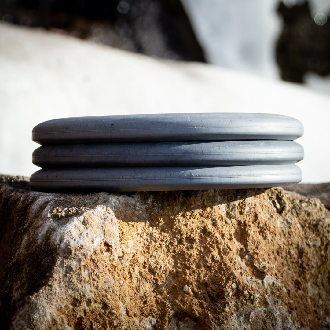 The Magic - Lunar, a round gray putter with three ridges, sits on a rough beige rock against a blurred snowy backdrop, ready for straight-flying glide.
