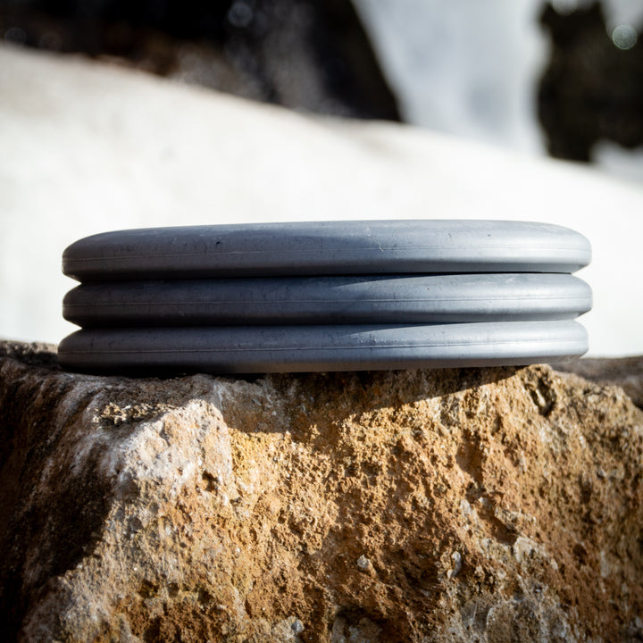 The Magic - Lunar, a round gray putter with three ridges, sits on a rough beige rock against a blurred snowy backdrop, ready for straight-flying glide.