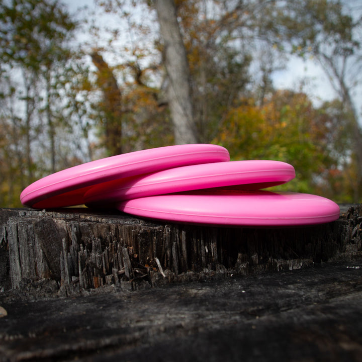 Three Magic - Money pink, beginner-friendly putters are stacked on a tree stump outdoors, framed by trees and autumn foliage in the background.