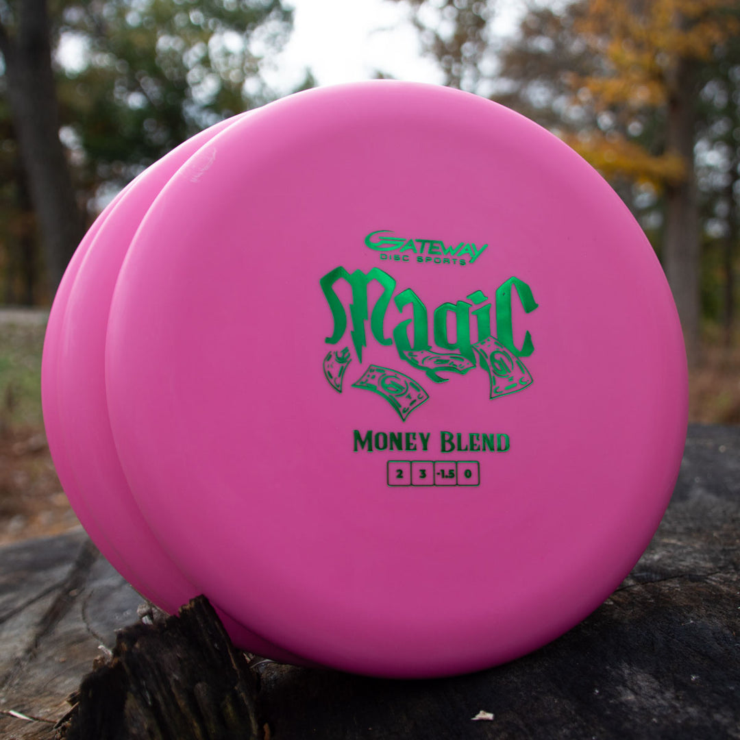 Two pink Gateway Magic - Money disc golf putters sit on a tree stump outdoors amid autumn trees. This straight-flying, beginner-friendly putter excels at accurate approaches, making it ideal for any disc golf course.