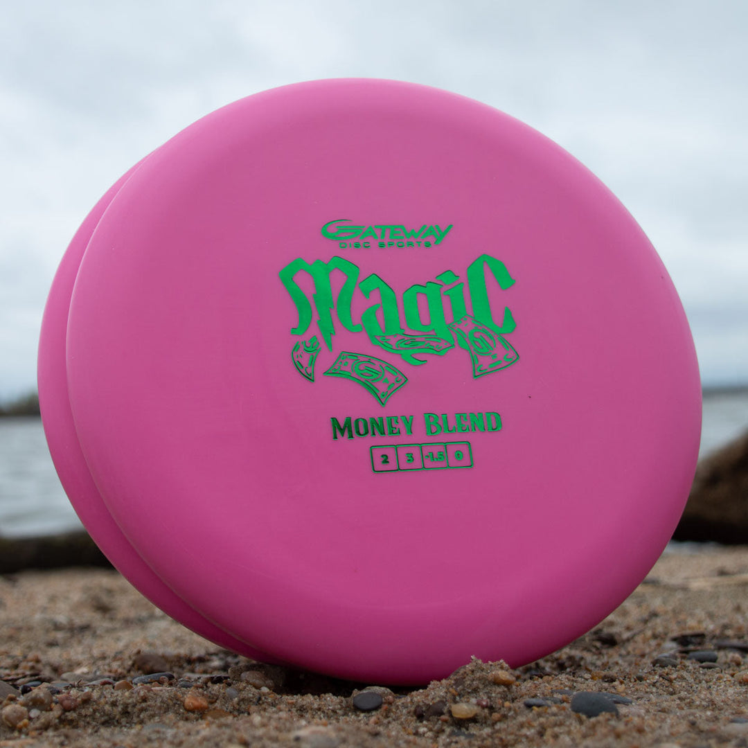 A pink Magic - Money beginner-friendly putter stands upright on sandy ground by the water, set for straight-flying putts and approach shots.