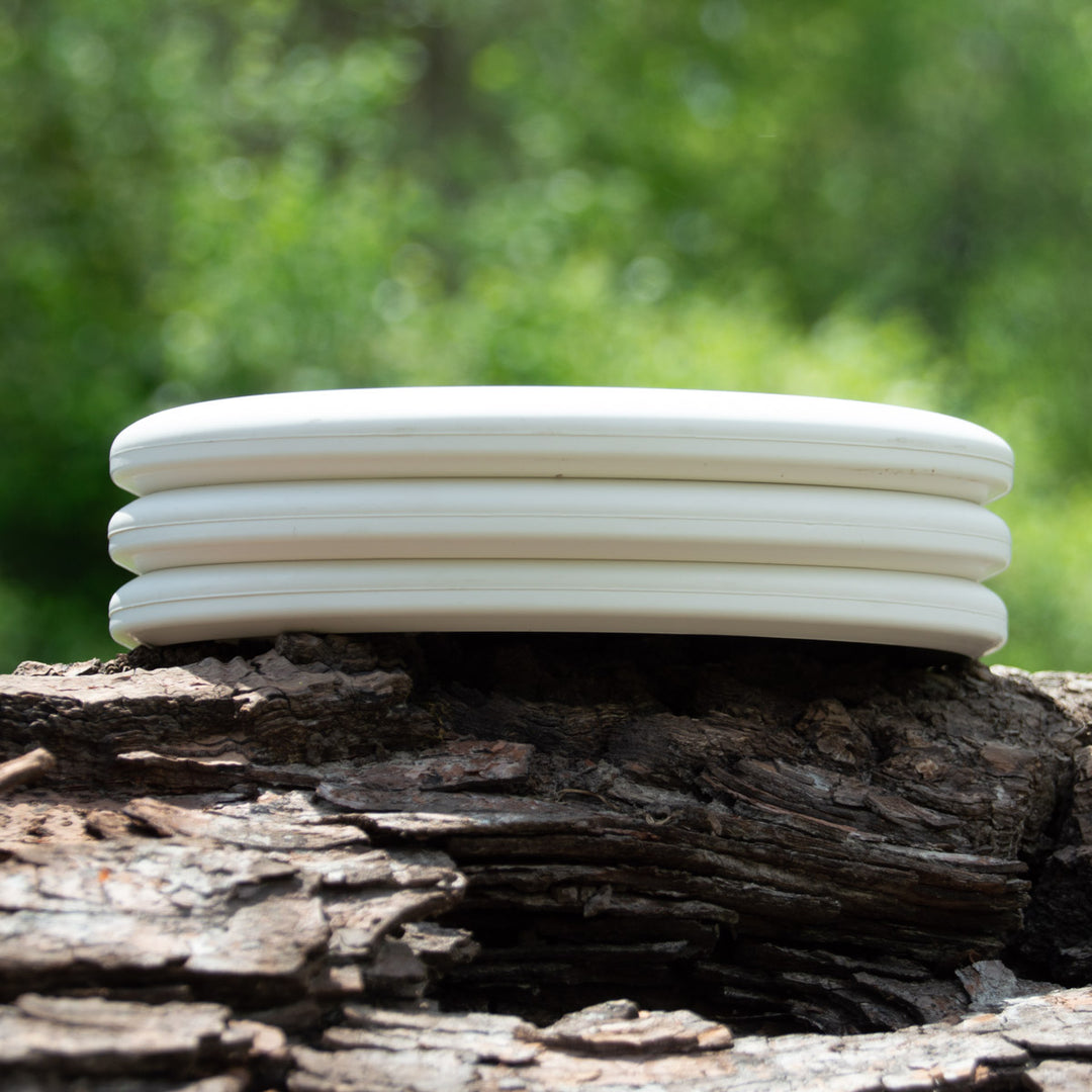 The Magic - Pure White beginner discs are stacked on rough tree bark, set against a blurred green background.