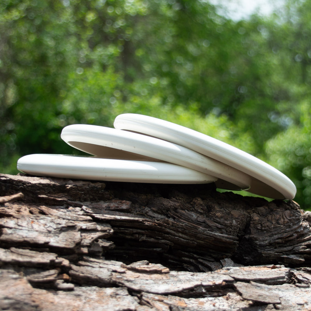 Three Magic - Pure White beginner discs, perfect for putt and approach shots, are stacked on a rugged tree log outdoors with blurred green foliage in the background.