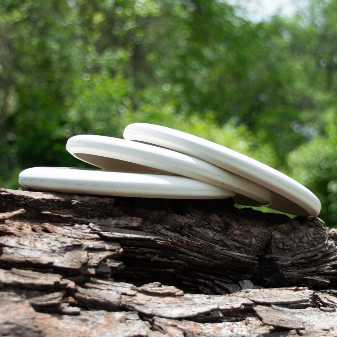 Three Magic - Pure White beginner discs, perfect for putt and approach shots, are stacked on a rugged tree log outdoors with blurred green foliage in the background.