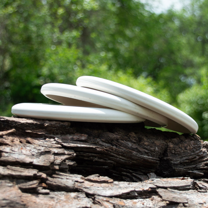 Three Magic - Pure White beginner discs, perfect for putt and approach shots, are stacked on a rugged tree log outdoors with blurred green foliage in the background.