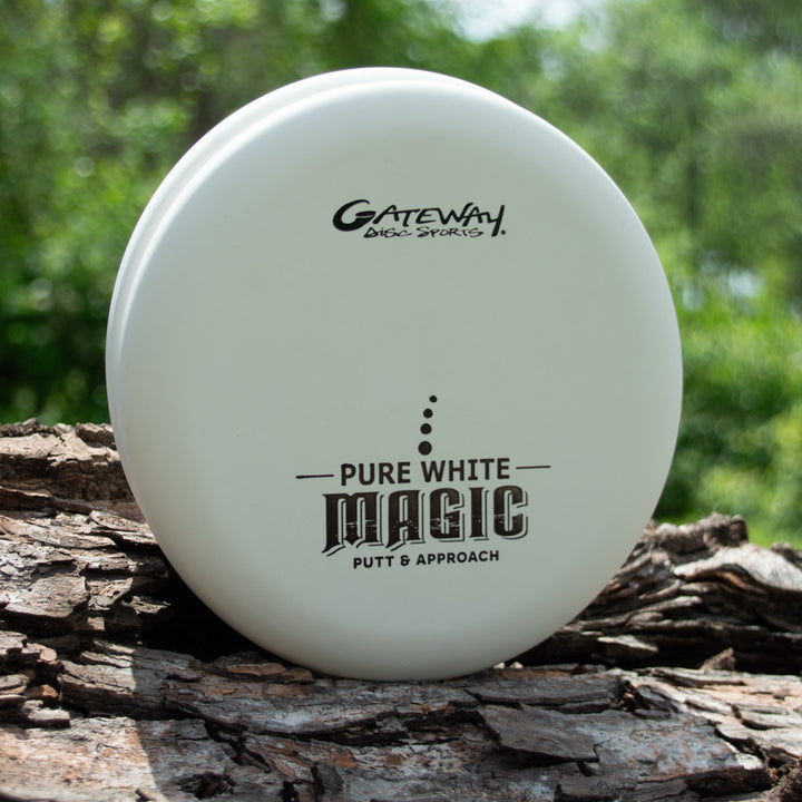 A Pure White Magic putt and approach disc by Gateway Disc Sports, a straight-flying putter ideal for beginners, stands upright on bark with greenery behind it.