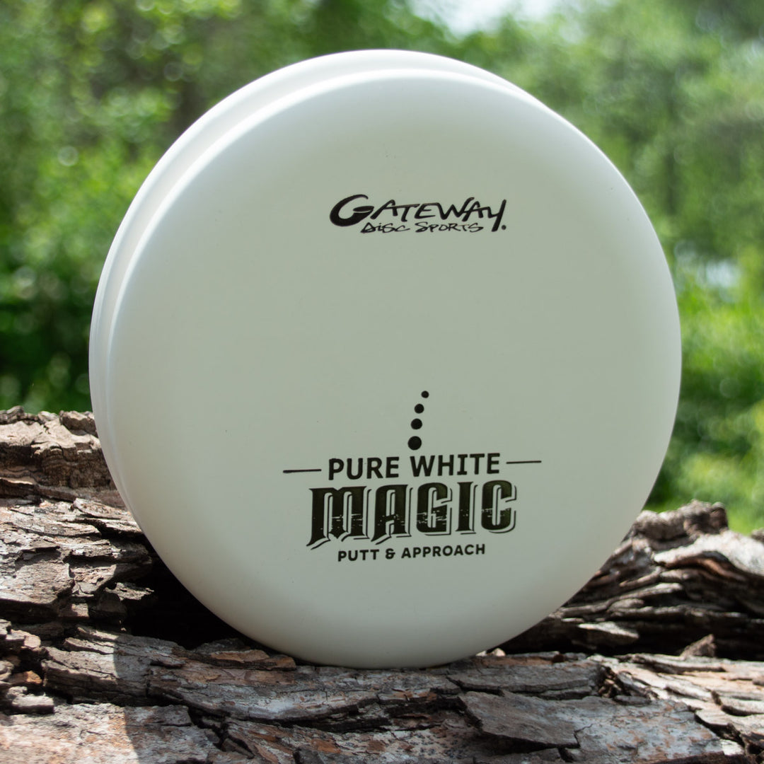 The straight-flying Magic - Pure White putter rests on a tree trunk, surrounded by greenery in the background.
