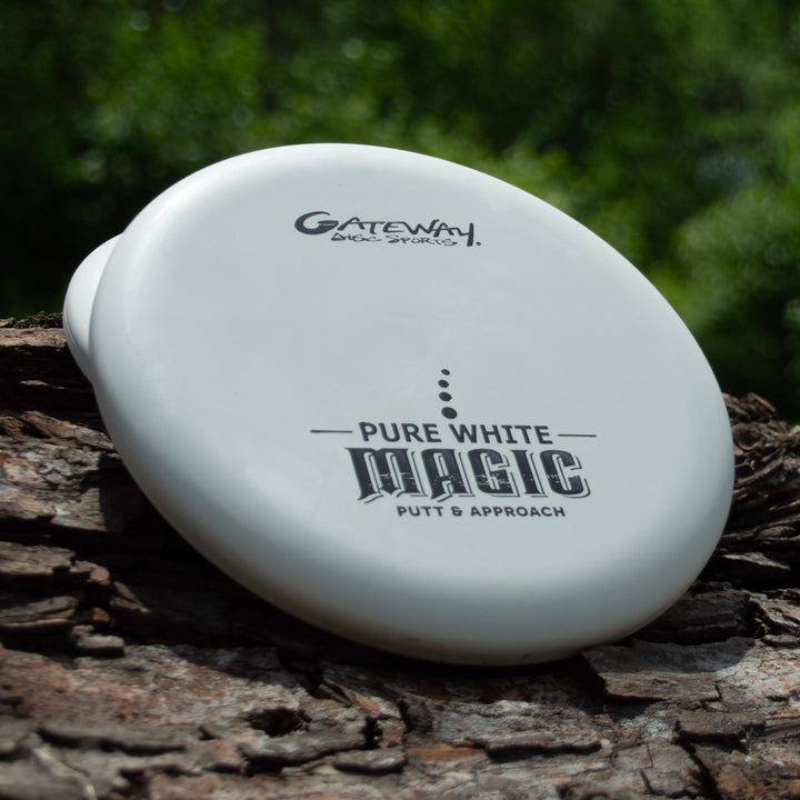 A white, straight-flying putter called "Magic - Pure White" rests on a tree trunk with green foliage in the background.