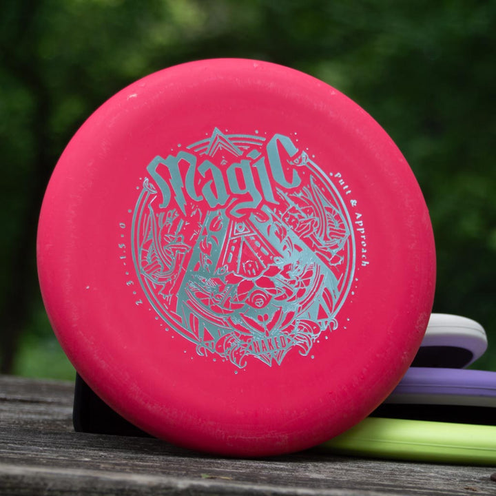 A pink Magic - Suregrip NAKED disc golf putter rests on a wooden surface outdoors, featuring reliable glide and straight-flight performance against a blurred green backdrop.