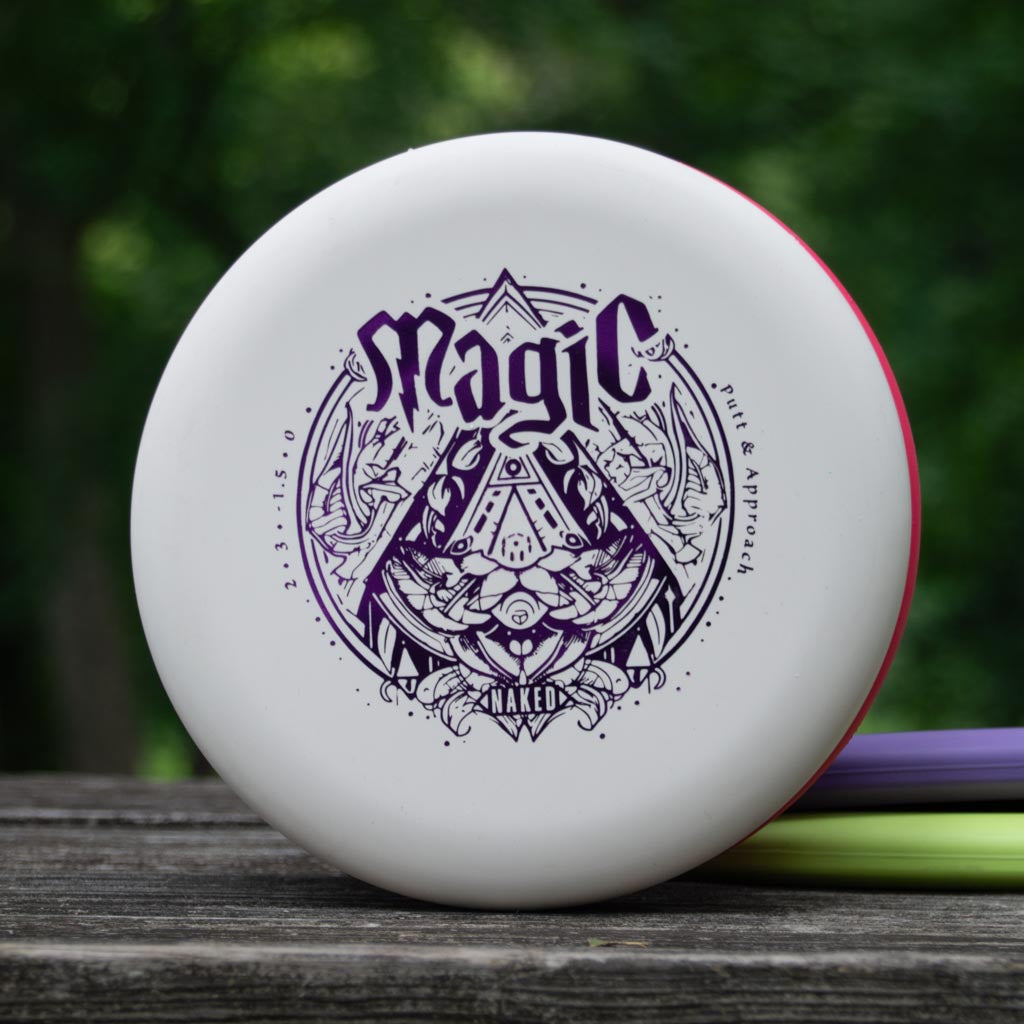 A white Magic - Suregrip NAKED disc golf putter with detailed purple graphics lies on a wooden surface, set against a blurred green outdoor scene.