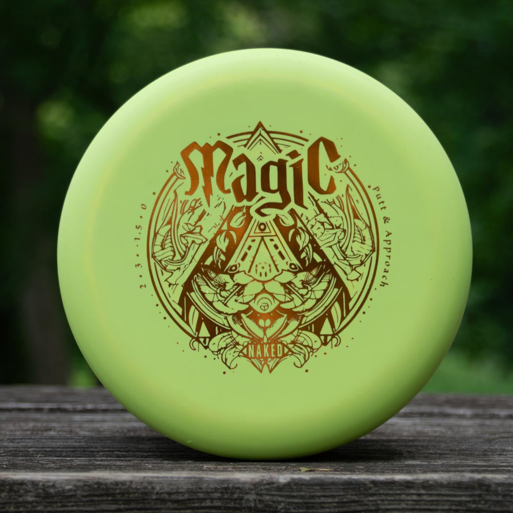 A light green Magic — Suregrip NAKED disc golf putter with detailed brown artwork and text sits on a wooden surface against a blurred green background, ready to display its straight flight and impressive glide.