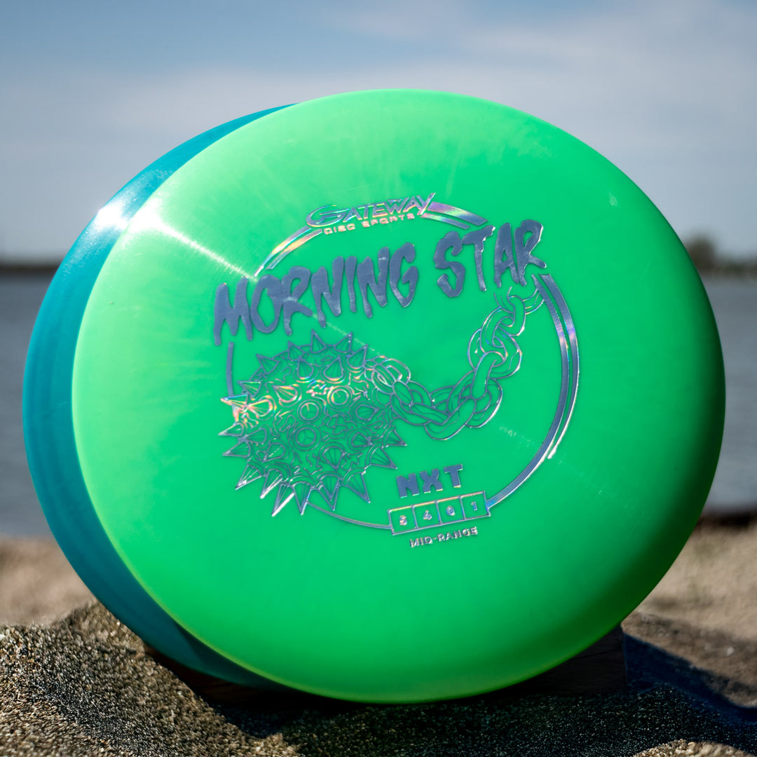 A Morningstar - NXT disc golf disc, green with a "Morning Star" label and silver chain-spiked design, features a neutral mid-range flight. It's shown on sand by water under a blue sky.