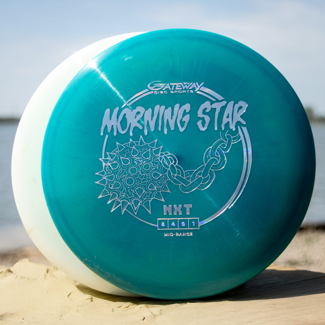 A teal and white "Morningstar - NXT" disc golf mid-range by Gateway Disc Sports, molded in durable NXT plastic, rests on sand near water - ideal for those seeking a neutral flying mid-range.