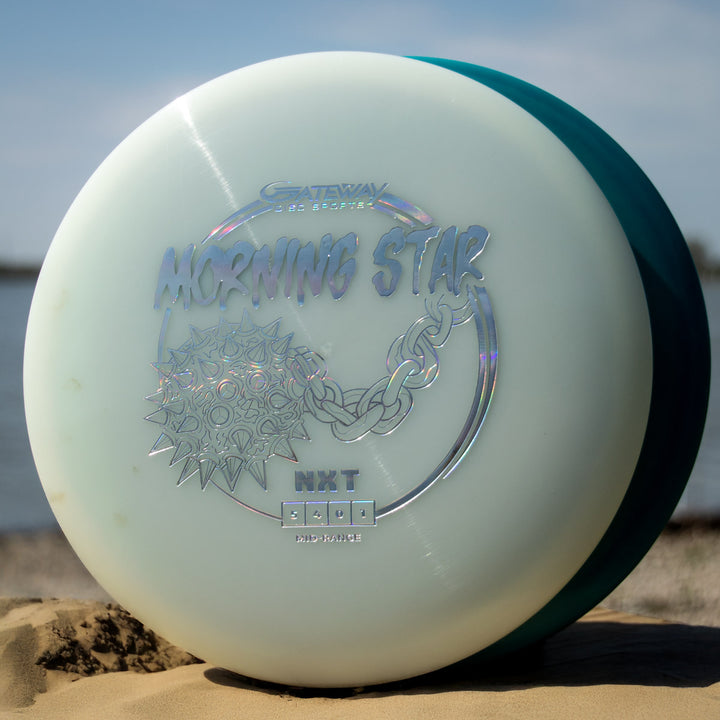 A close-up of a pale blue Morningstar - NXT disc golf mid-range with metallic lettering, standing upright on sandy ground by water - a reliable, neutral-flying mid-range in durable NXT plastic.