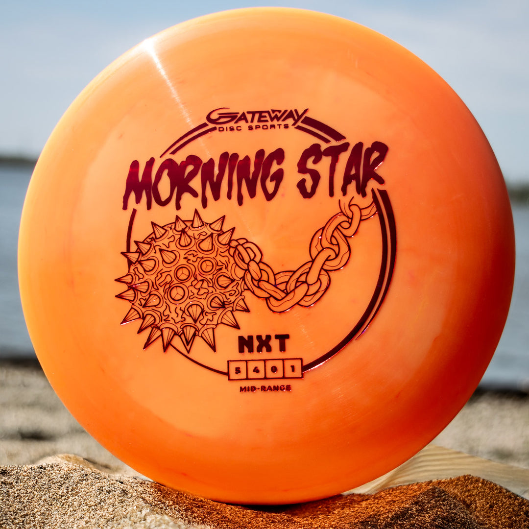 An orange Morningstar - NXT mid-range disc from Gateway Disc Sports, with Suregrip molds, lies on sandy ground by the water.