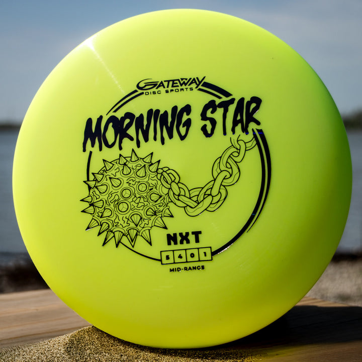 A yellow Morningstar - NXT mid-range disc with a spiked mace and chain graphic sits on sand outdoors. Made with durable NXT plastic, this neutral flying disc offers consistent performance for disc golf.