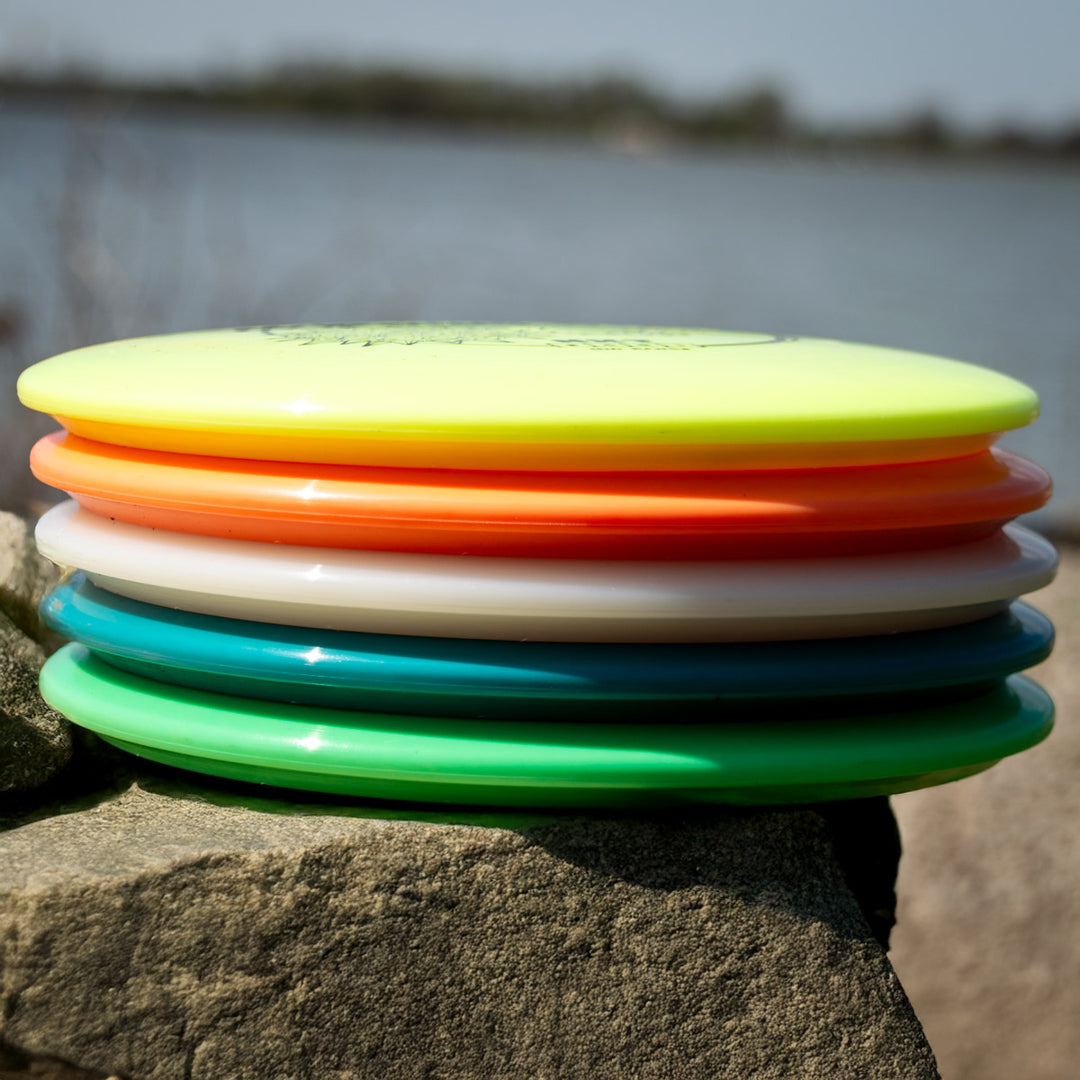 A stack of colorful Suregrip molds sits on a rock near the water with trees in the background is perfect for testing your Morningstar - NXT or any neutral flying mid-range.