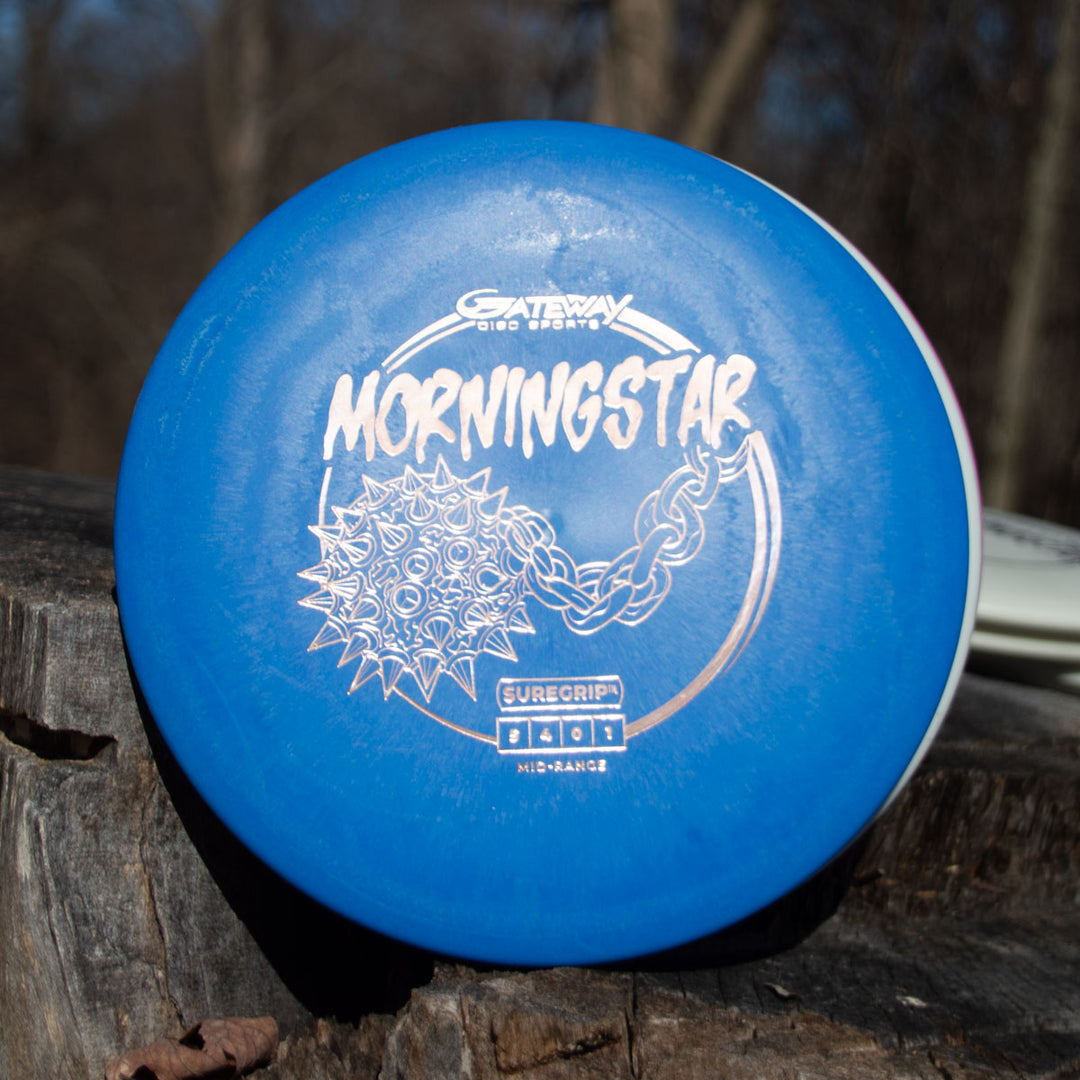 A blue Gateway Morningstar - Suregrip disc golf disc with a spiked ball and chain graphic sits on a tree stump. This neutral-flying mid-range is ideal for versatile throws.