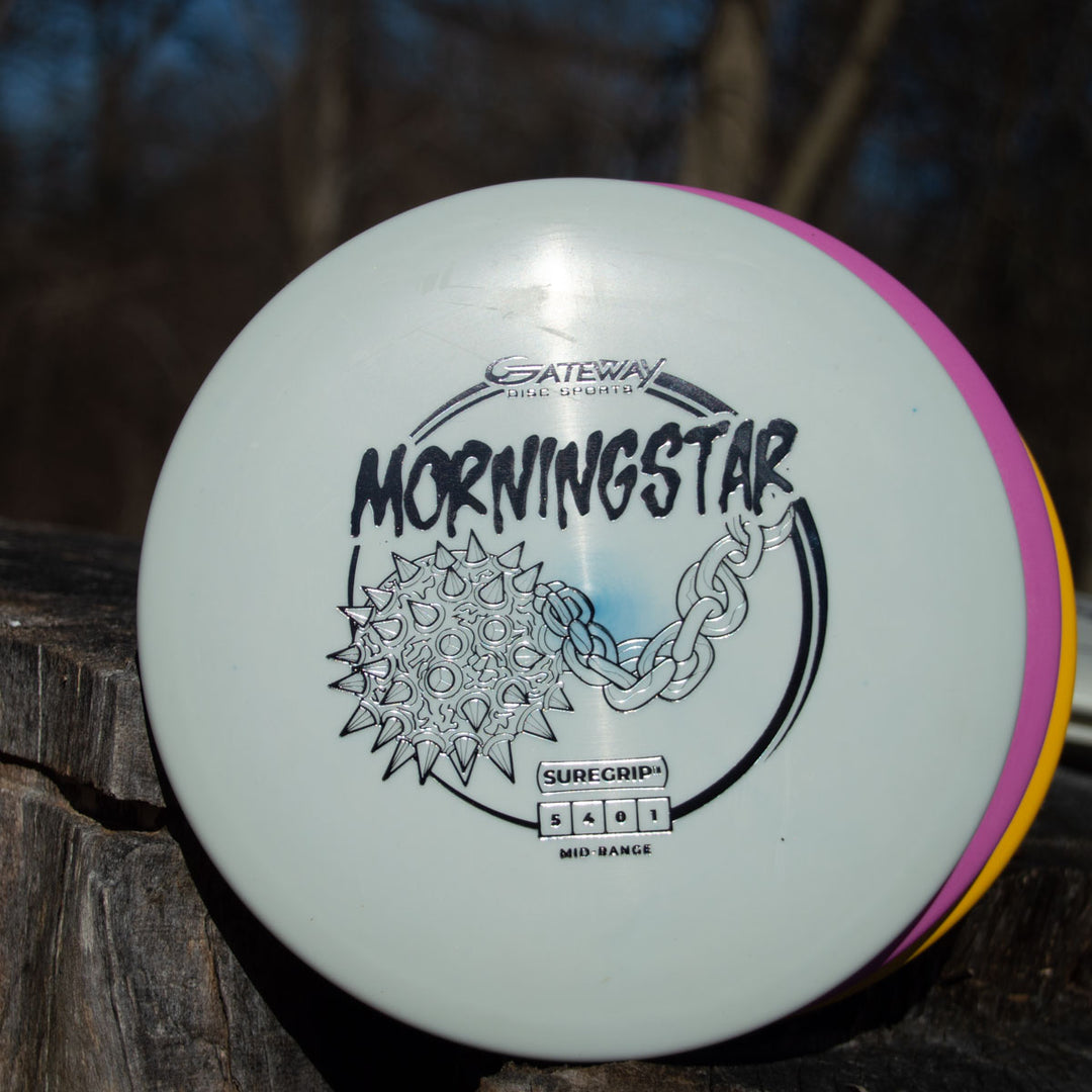 A white Morningstar - Suregrip disc golf disc with a spiked ball and chain design sits on wood outdoors. This neutral flying mid-range is crafted from Suregrip plastic for reliable throws.