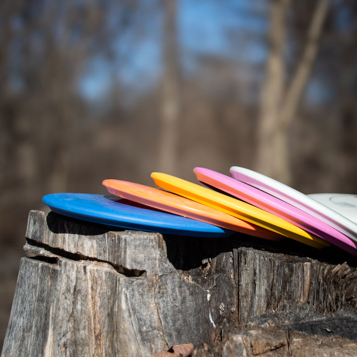 Five colorful disc golf discs, including the Morningstar - Suregrip neutral-flying mid-range, are stacked on a weathered tree stump outdoors with blurred leafless trees in the background.