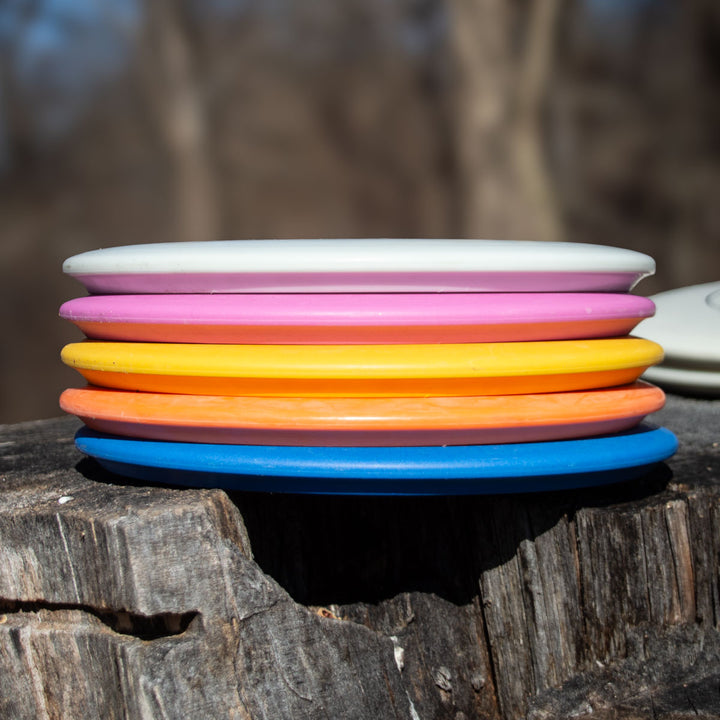 A stack of five colorful Morningstar - Suregrip frisbees in white, pink, orange, yellow, and blue rests on a tree stump outdoors with a blurred background. Ideal for fans of this neutral flying mid-range disc.