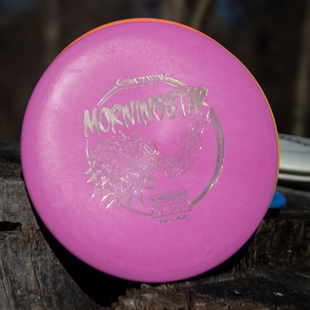 A pink Morningstar - Suregrip neutral flying mid-range disc rests on a tree stump, its plastic gleaming in sunlight against a blurred natural background.