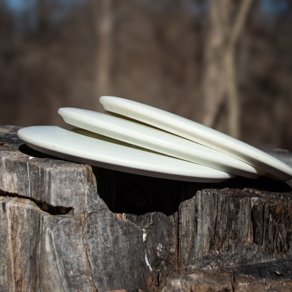 Three white Morningstar - Suregrip Superglow molds are stacked on a tree stump against a blurred forest backdrop—ideal for anyone seeking a neutral flying mid-range.