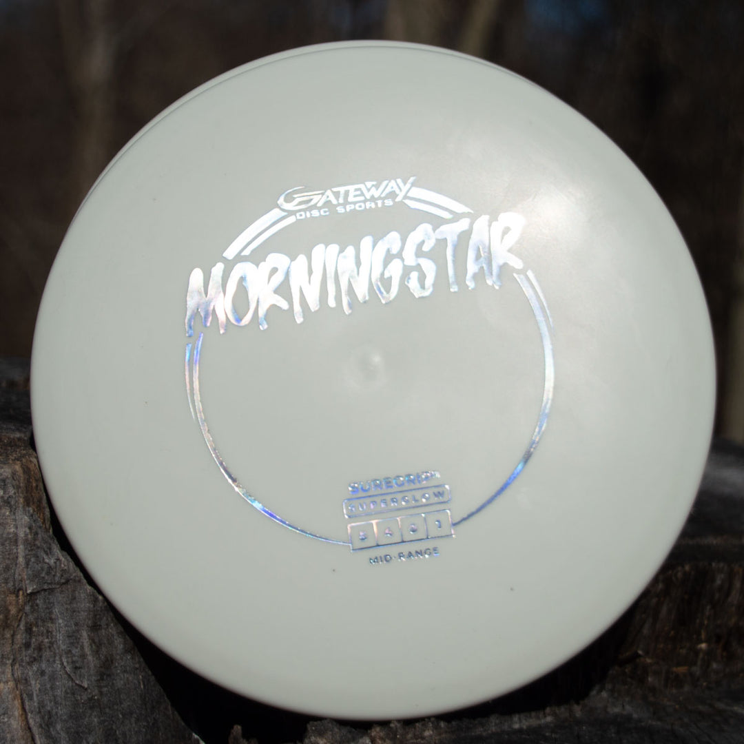 A white Morningstar - Suregrip Superglow mid-range disc from Gateway Disc Sports rests on weathered wood outdoors, silver lettering visible—a dependable, neutral-flying disc that showcases the brand's Suregrip molds.
