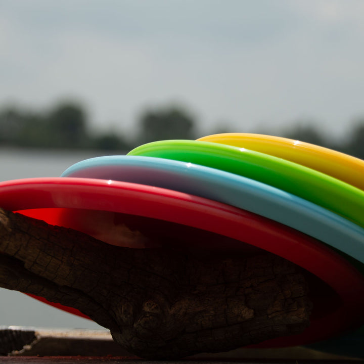 The Mystic - NXT, a colorful plastic mid-range disc recognized for its glide and straight flight, rests on a wooden surface outdoors with blurred trees and water in the background.