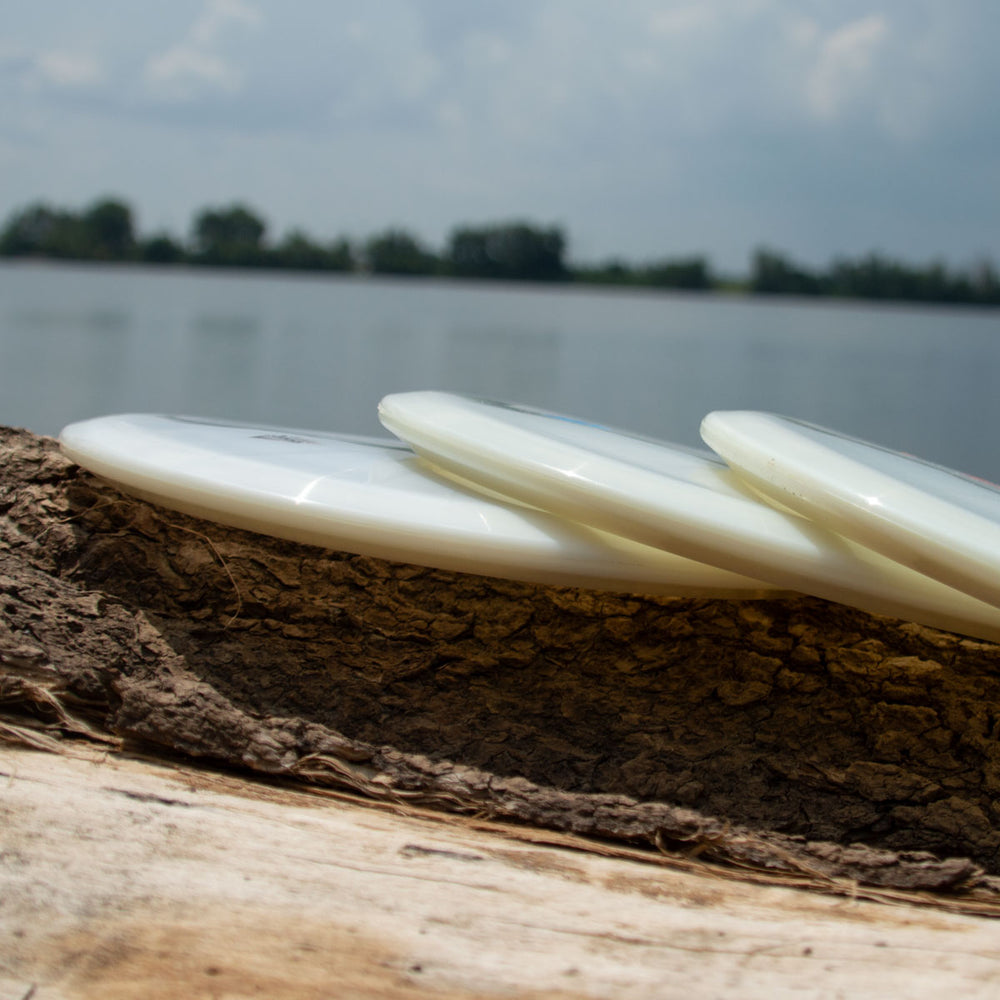 Three Mystic - Diamond Superglow white glow-in-the-dark frisbees are stacked on a log by the water, with trees and a cloudy sky behind them.