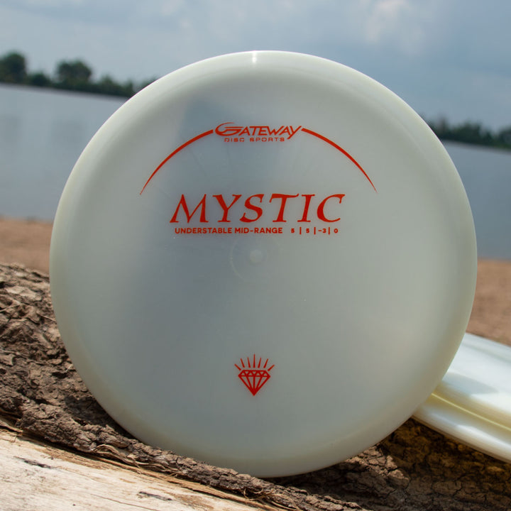 A white Mystic - Diamond Superglow disc golf disc with orange text, valued for its controllable mid-range and straight flight, rests on a log near a lake and sandy shore.
