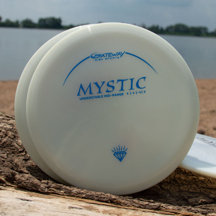 A white Mystic - Diamond Superglow mid-range disc golf disc from Gateway Disc Sports sits on a log at a beach, with water and trees in the background.