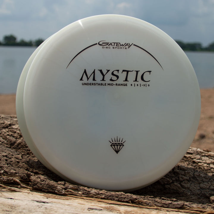 A white Gateway Mystic - Diamond Superglow disc golf disc labeled "UNDERSTABLE MID-RANGE" rests on wood by water, known for its controllable mid-range and straight flight.