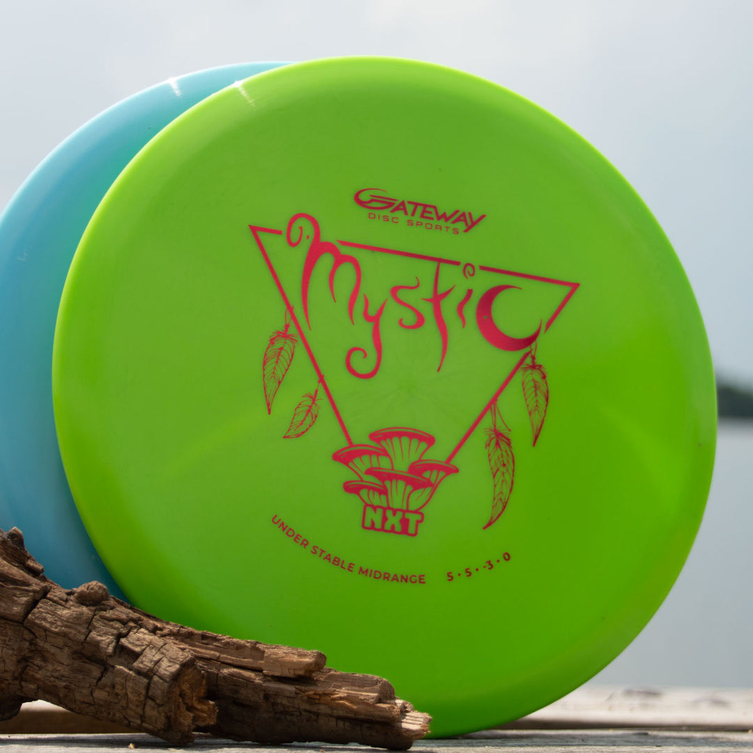 The lime green Mystic - NXT mid-range disc by Gateway Disc Sports, praised for its straight flight and glide, features a pink design and rests against a blue disc on a wooden outdoor surface.