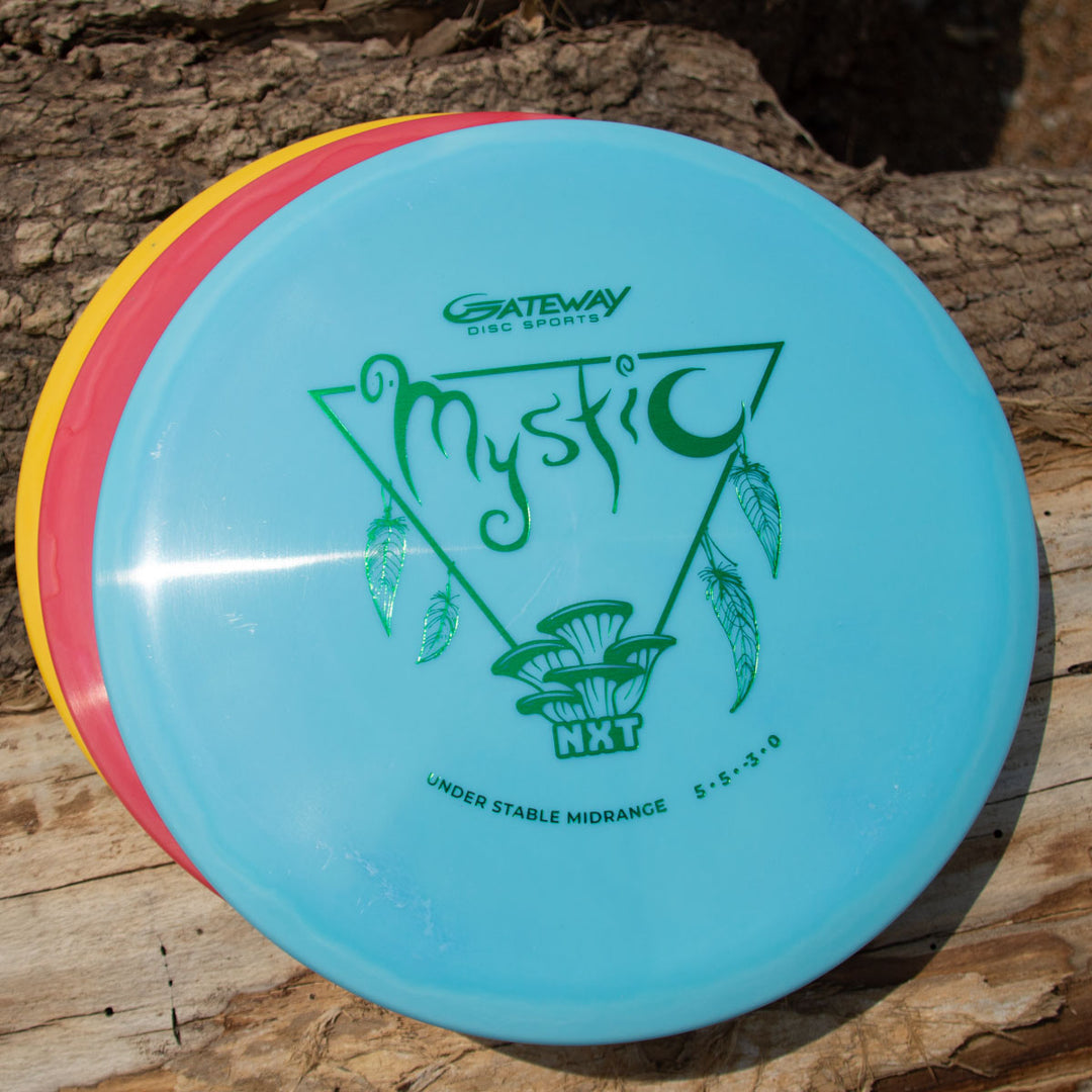 Three disc golf discs—yellow, red, and a blue mid-range disc labeled "Mystic - NXT" with green graphics—are stacked on a wooden surface. The Mystic - NXT is known for its excellent glide and straight flight.