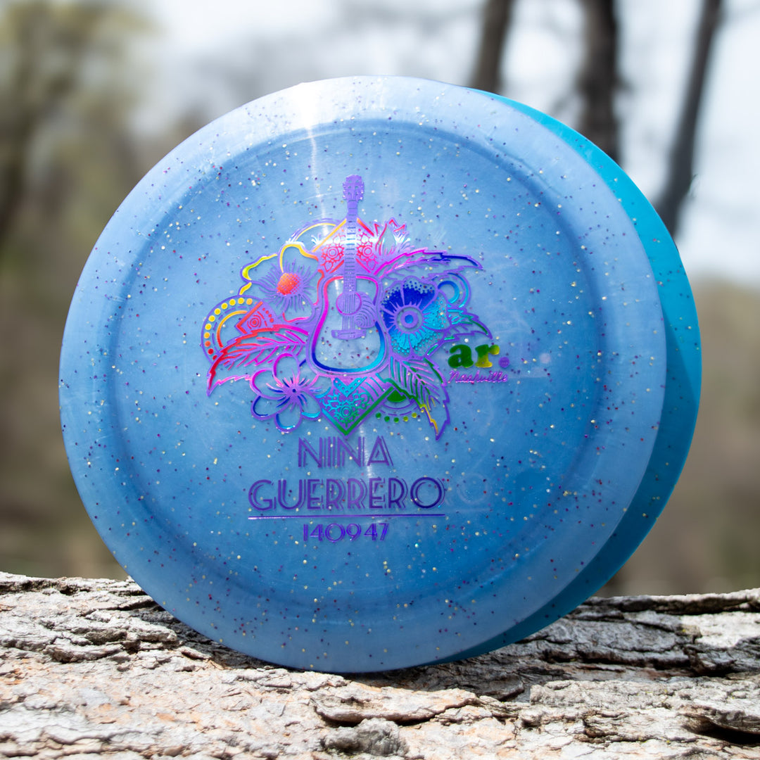 Nina Guerrero's Aura - Diamond Metal Flake: A blue, glittery disc golf disc with a vibrant guitar/floral design and "Nina Guerrero 140947" printed on it, displayed on a tree branch—ideal for stylish throws with great turnover ability.