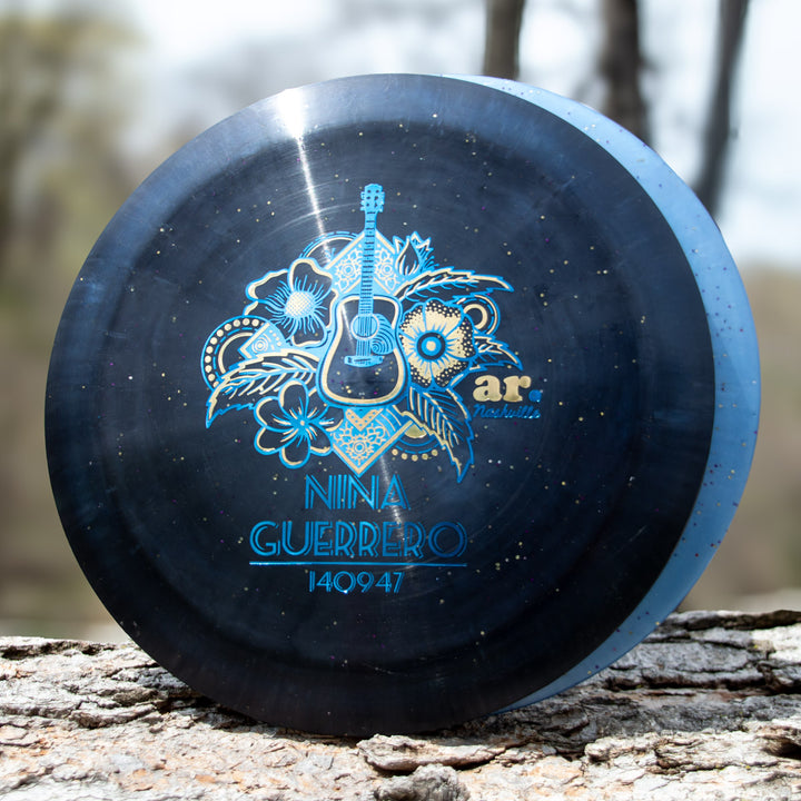 A round, dark blue disc named "Nina Guerrero's Aura - Diamond Metal Flake," with floral and guitar art plus "Nina Guerrero 140947," "ar Nashville," and Flight Numbers, rests on a tree log outdoors.
