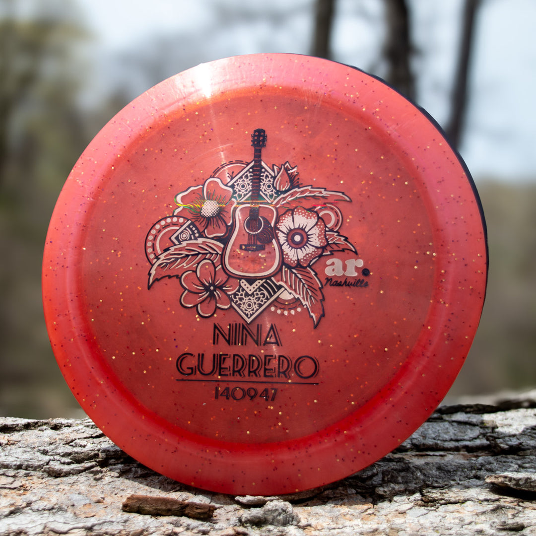 Nina Guerrero's Aura - Diamond Metal Flake, a red speckled disc golf disc with floral and guitar art, sits on tree bark outside.