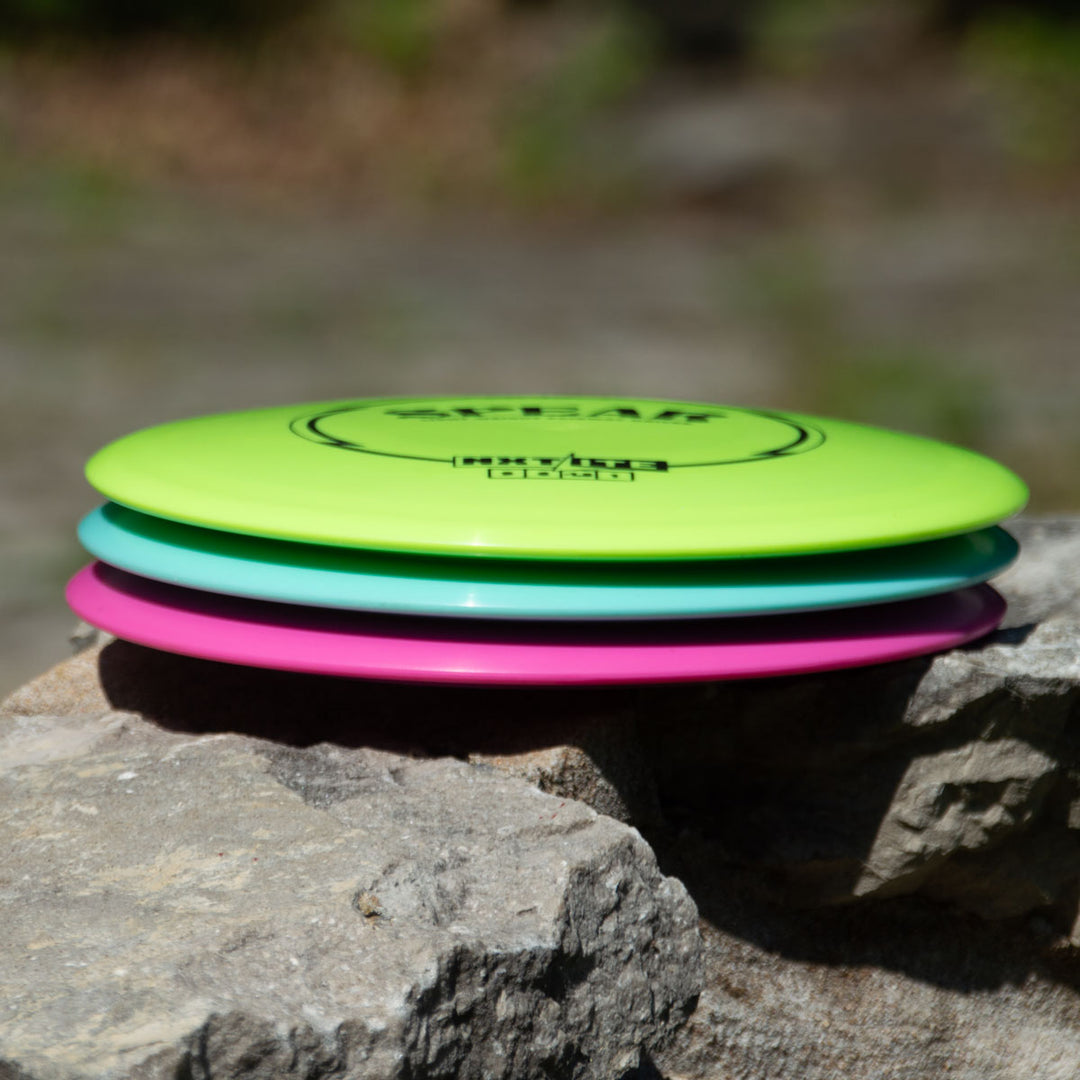 The Spear - NXT LITE, available in vibrant green, blue, and pink, is shown stacked on a rock outdoors with a blurred natural background—perfect for players seeking accuracy on the disc golf course.