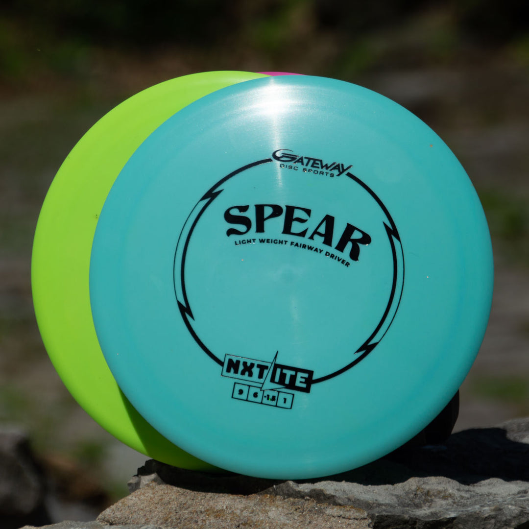 Two disc golf discs, one blue and one yellow, rest upright against a rock. The blue disc shows "Spear - NXT LITE" from Gateway Disc Sports, a control driver designed for precise placement on the course.