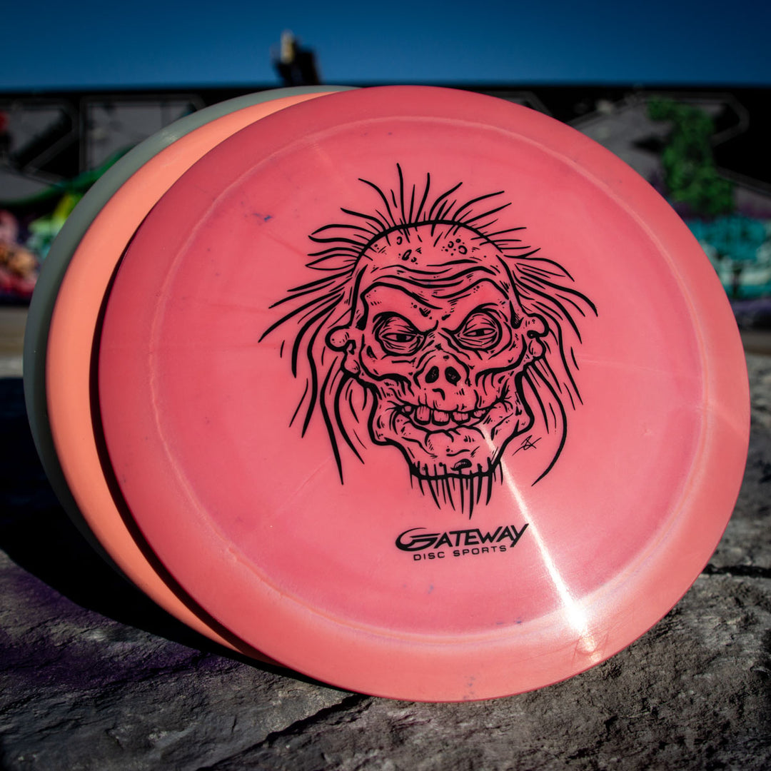 A pink Speed Demon - Nylon - Premier overstable distance driver with a black zombie-like face illustration rests on a stone surface, ideal for powerful forehands or backhand flex shots.
