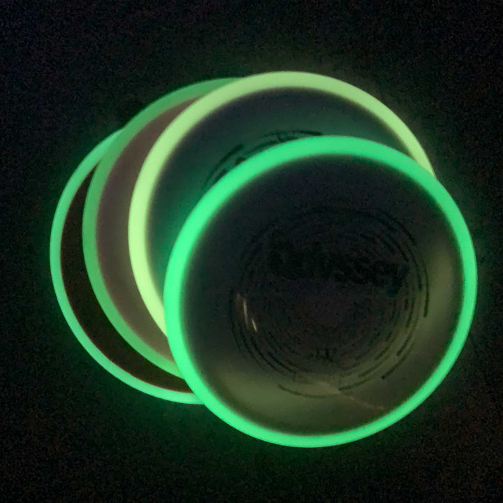 Three glowing green Odyssey - NXT Superglow discs, stacked slightly askew, are photographed in the dark—evoking the mysterious energy of an odyssey.