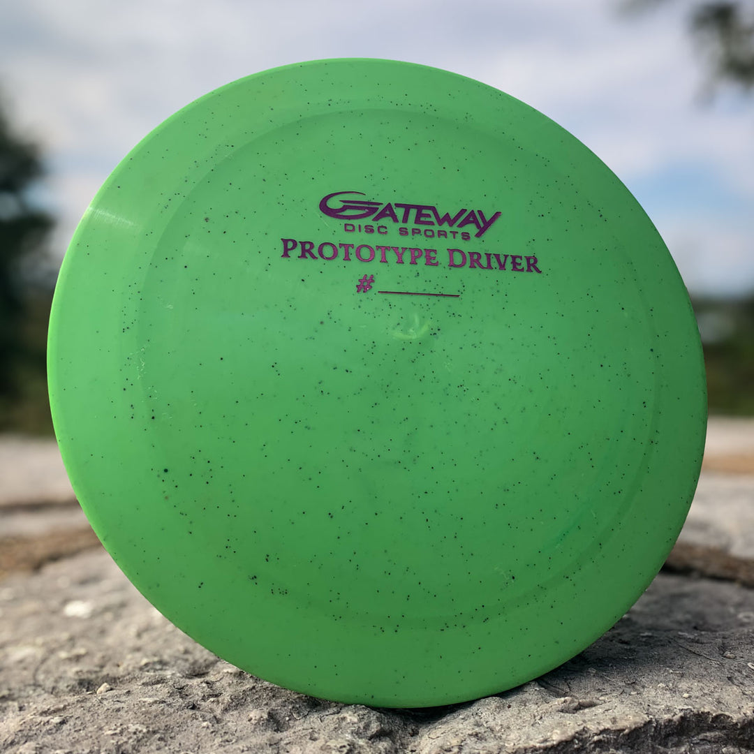 The Prototype 136 - NXT Hemp (Javelin) disc golf driver features a green color with black speckles and sits on a rock outdoors—ideal for players who want superior distance control.