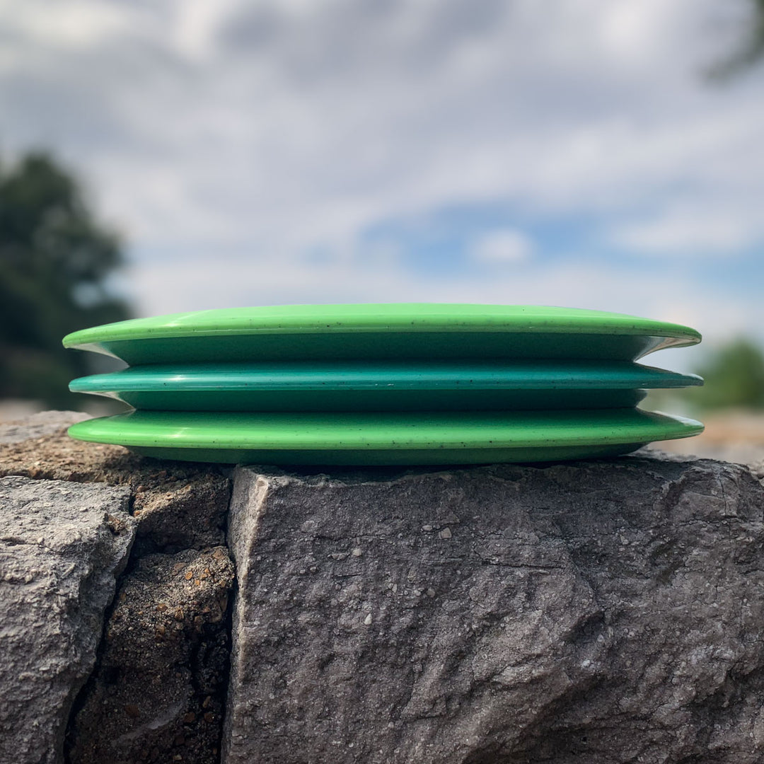 Three green Prototype 136 - NXT Hemp (Javelin) discs stacked on a large rock outdoors, set against a blurred natural background and cloudy sky, showcasing this trusted distance control driver.